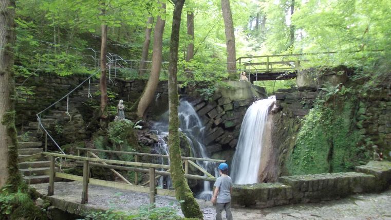 Ein kleiner Wasserfall in einem grünen Wald. Ein Junge steht auf einem gepflasterten Weg und beobachtet das Wasser.