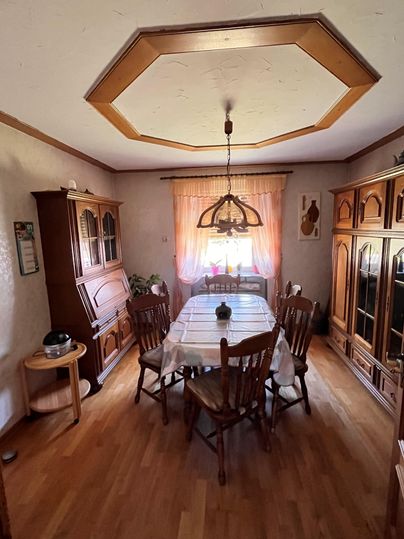 Een gezellige eethoek met een grote houten tafel en verschillende stoelen. Een raam met gordijnen zorgt voor natuurlijk licht en een warme sfeer.