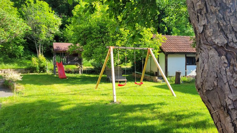 A playground with a swing and a slide in a green garden. In the background, there is a small house and many trees visible.