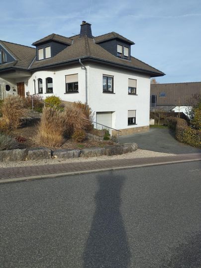 Ein modernes, zweigeschossiges Haus mit einem begrünten Vorgarten. Die Fassade ist weiß und das Dach ist dunkel.