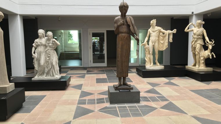 Un intérieur avec plusieurs sculptures en différents matériaux. Au centre se trouve une figure en bois, entourée de statues en marbre.