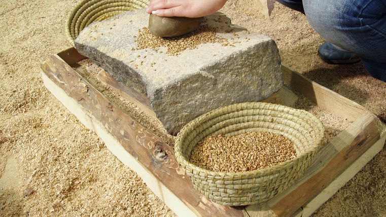 Eine Person malt Getreide mit einem Stein auf einem flachen Stein. Daneben sind Körbe mit gemahlenem Getreide platziert.
