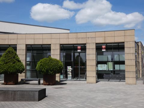 Ein modernes Gebäude mit großen Glasfenstern und einer zeitgenössischen Architektur. Vor dem Gebäude befinden sich gepflegte Pflanzen und eine gepflasterte Fläche.