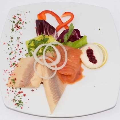 Ein ansprechend angerichteter Fischteller mit verschiedenen Fischsorten, frischen Salatblättern und einer Zitronenscheibe. Dazu sind rote Paprikastreifen und eine kleine Portion Sauce dekoriert.