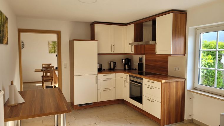 A bright, modern kitchen with wooden and white elements. In the background, a dining area can be seen.