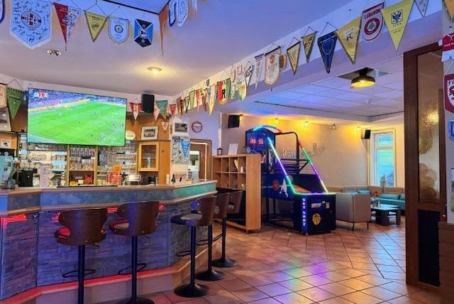 A cozy interior of an establishment with a bar, seating, and a large television screen for sports broadcasts. Numerous flags hang on the walls and there is a jukebox in the corner.