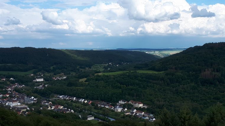 Une vaste vue sur des collines vertes et un petit village. Le ciel est nuageux avec quelques zones bleues.