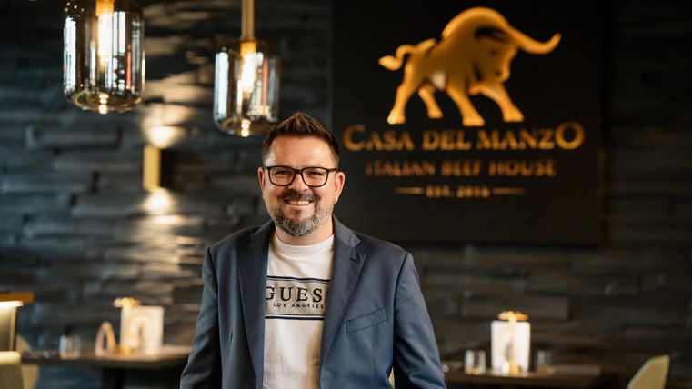 Un homme souriant se tient dans un restaurant élégant avec l'inscription "Casa del Manzo" en arrière-plan. L'ambiance est moderne et accueillante.
