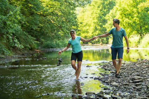Ein Paar spaziert barfuß durch einen flachen Fluss, umgeben von grüner Natur.
