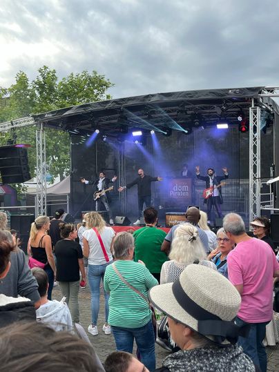 A band is playing on a stage in front of an enthusiastic audience. Many people are gathered together, enjoying the music under a cloudy sky.
