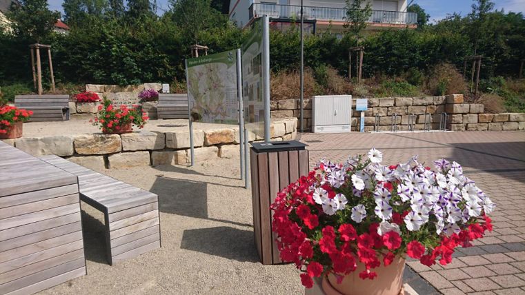Ein hübscher Außenbereich mit Holzsitzen und bunten Blumen. Im Hintergrund steht ein Informationsschild und grüne Sträucher umgeben die Atmosphäre.