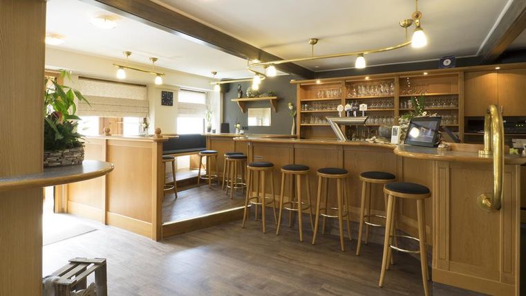 Eine gemütliche Bar mit Holzmöbeln und Barhockern. Die Atmosphäre ist einladend und hell, mit Pflanzen und einer gut sortierten Theke.