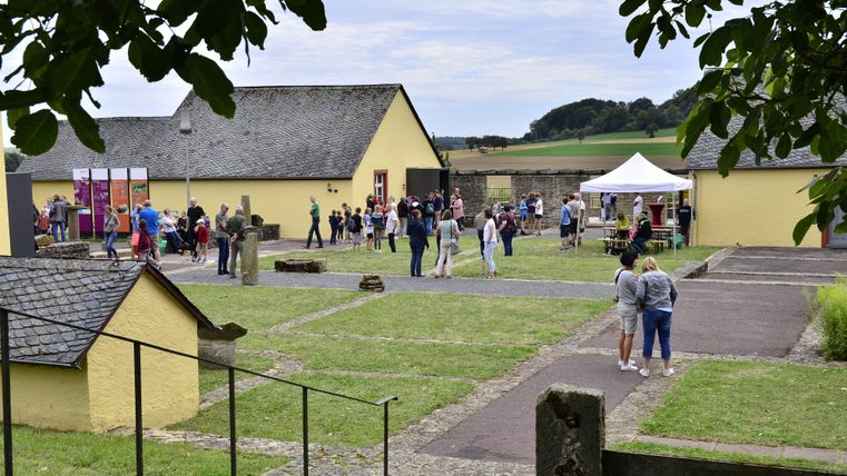 A group of people gathers on a spacious area with yellow buildings. In the background, tents and a green landscape are visible.