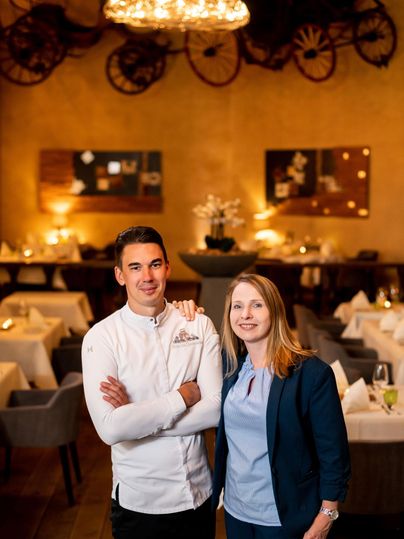 A restaurant with elegantly decorated tables and warm lighting. Two people are smiling in the foreground.