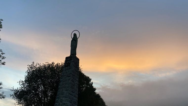 A statue stands on a high stone with a gentle, colorful sky in the background. Trees surround the statue and it is slightly foggy.