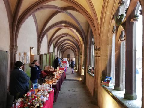 Un couloir historique avec des arcs gothiques. Aux murs, il y a des tables avec différents produits artisanaux et les visiteurs explorent les stands.