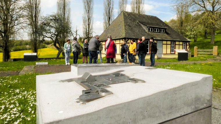 Un groupe de personnes se rassemble devant un bâtiment historique dans un paysage verdoyant. Au premier plan, on voit un monument gris.