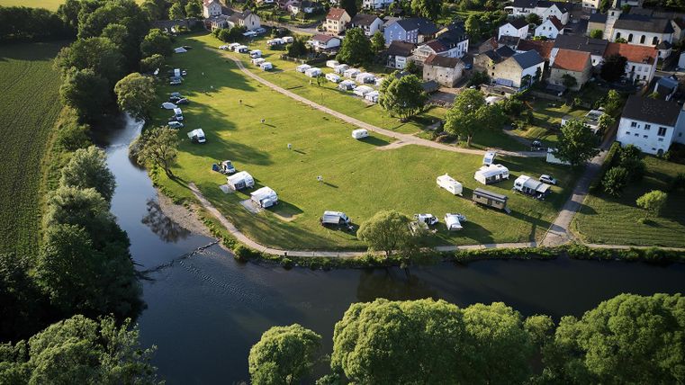 A campsite by the river with many RVs and tents. In the background, houses and a green landscape can be seen.