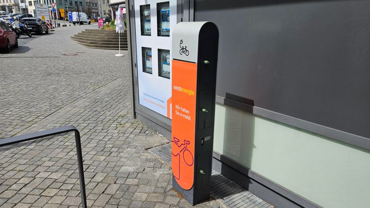 Une station de vélo avec un panneau d'information orange est située sur un chemin pavé. En arrière-plan, plusieurs bâtiments et des voitures garées sont visibles.