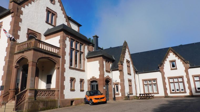 Ein historisches Gebäude mit einer charmanten Fassade. Vor dem Gebäude steht ein orangefarbenes Auto auf einem gepflasterten Platz.