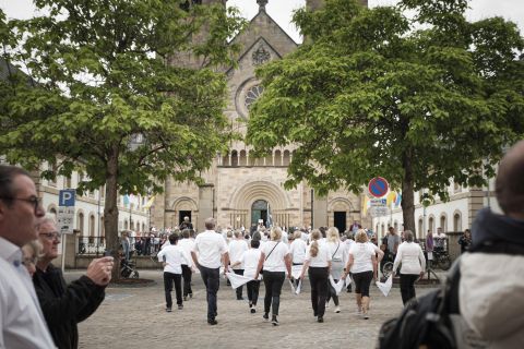 Une grande foule se rassemble sur une place. Une parade avec des musiciens en uniformes rouges et blancs passe.