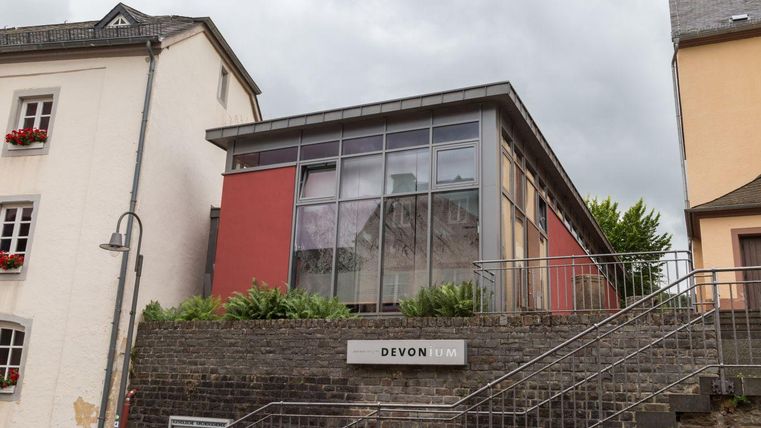 A modern building with large windows and a red facade. It is located at a staircase and is surrounded by green plants.
