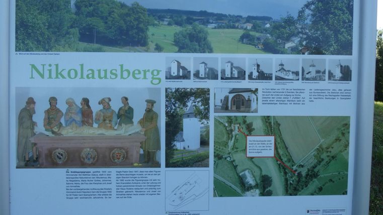 Eine Infotafel über den Nikolausberg mit Bildern und Informationen zu den Sehenswürdigkeiten. Die Landschaft im Hintergrund ist grün und hügelig.