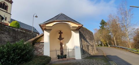 Ein einfacher Kreuzweg mit einer Statue in einer Nische. Im Hintergrund sind Bäume und ein klarer Himmel zu sehen.