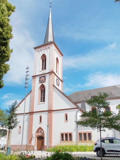 Een mooie kerk met een hoge toren en een blauwe lucht. Rondom de kerk zijn er bomen en een parkeerplaats.