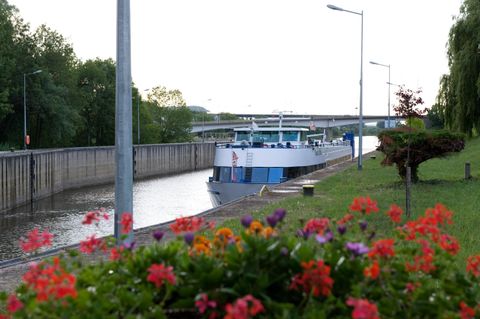 Een schip vaart door een kanaal, omgeven door een groene landschap. Op de voorgrond bloeien kleurvolle bloemen.