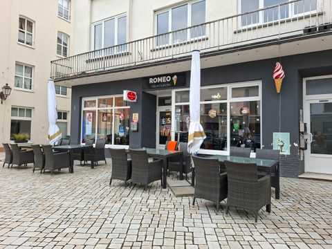 Un café avec des places en extérieur et des parasols. Le bâtiment a de grandes fenêtres et une atmosphère accueillante.
