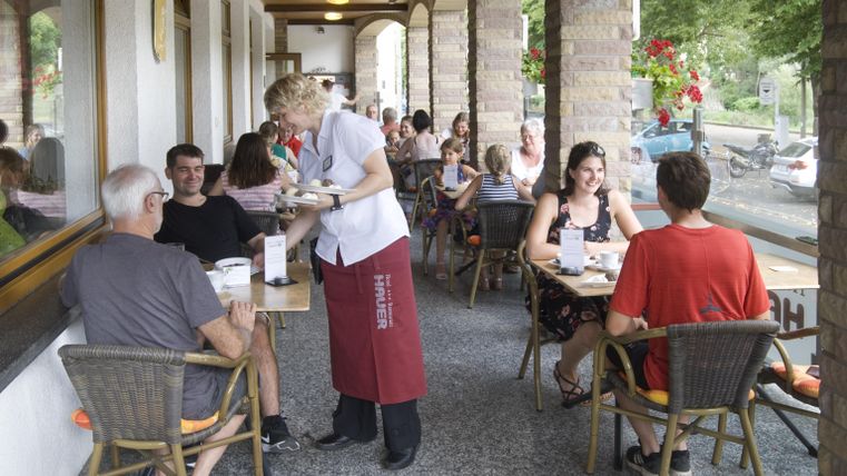 Eine lebhafte Terrasse eines Restaurants mit Gästen, die an Tischen sitzen. Eine Bedienung serviert Speisen und das Ambiente ist freundlich und entspannt.