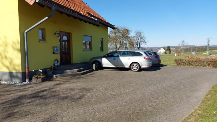 A yellow house with a bright sky and a paved front yard. A silver car is parked in front of the entrance.