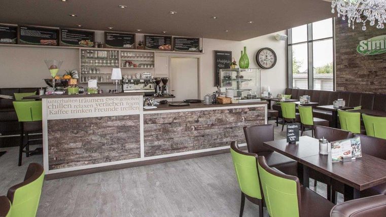 Ein modernes Café mit Holz- und Grüntönen. Tische und Stühle bieten eine gemütliche Atmosphäre, während eine Theke mit verschiedenen Getränken und Snacks im Hintergrund sichtbar ist.