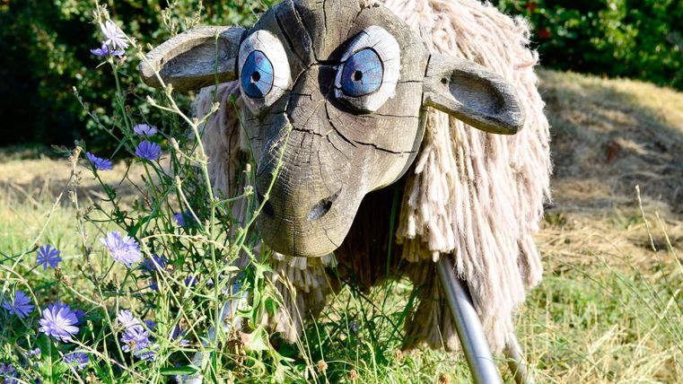 Een decoratieve houten schapen met een grappige mimiek staat in het gras. Het is omgeven door kleurrijke bloemen en groene vegetatie.