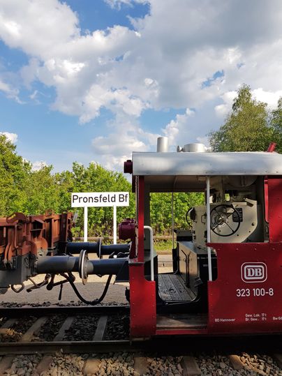 A red train is standing on the tracks at the Pronsfeld Bf station. The sky is partly cloudy with some nice clouds.