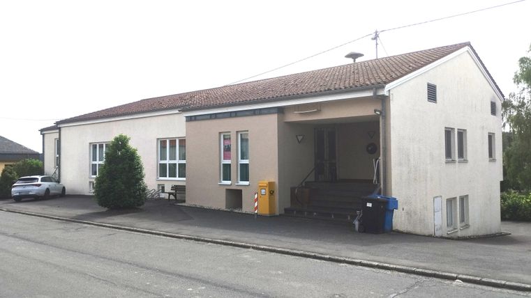 Un bâtiment simple avec une façade blanche et un toit rouge. Devant le bâtiment, il y a quelques arbres et des voitures garées.