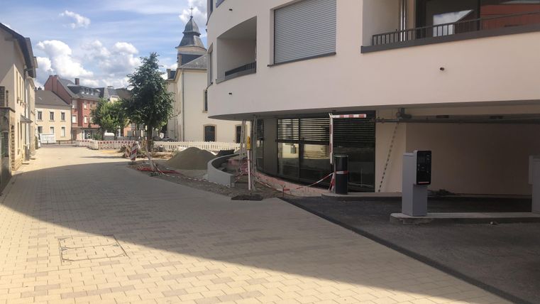 Eine ruhige Straße mit modernen Gebäuden und viel Tageslicht. Auf der linken Seite sind Bäume und Baustellenmaterial sichtbar.