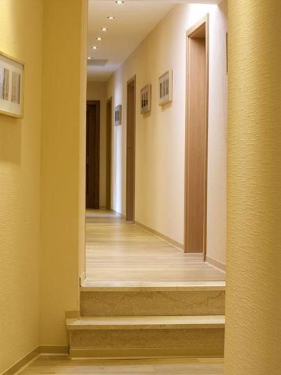 A bright hallway with wooden-colored flooring and soft beige walls. Some pictures hang on the walls, and the hallway leads to several doors.