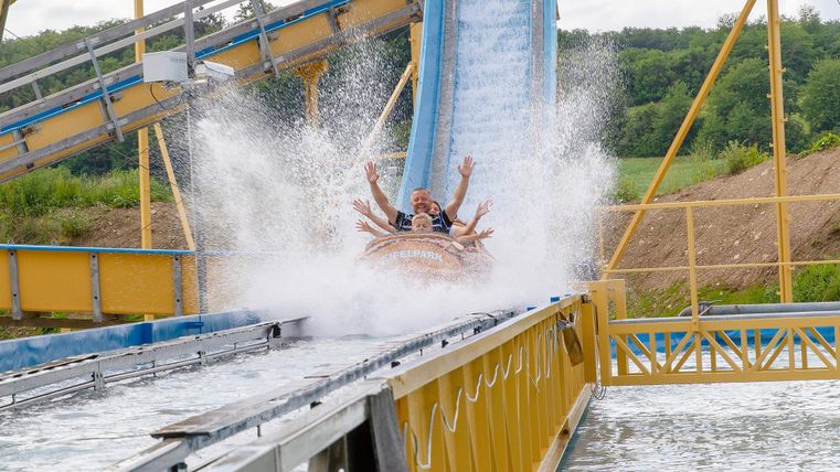 Eine aufregende Wasserbahn mit begeisterten Fahrgästen, die in eine Wasserfontäne rutschen. Im Hintergrund sind die Anlage und die ergrünte Landschaft zu sehen.