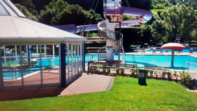 Ein Schwimmbad mit einer Rutsche und grünen Rasenflächen. Im Hintergrund sind Sonnenschirme und Bäume zu sehen.