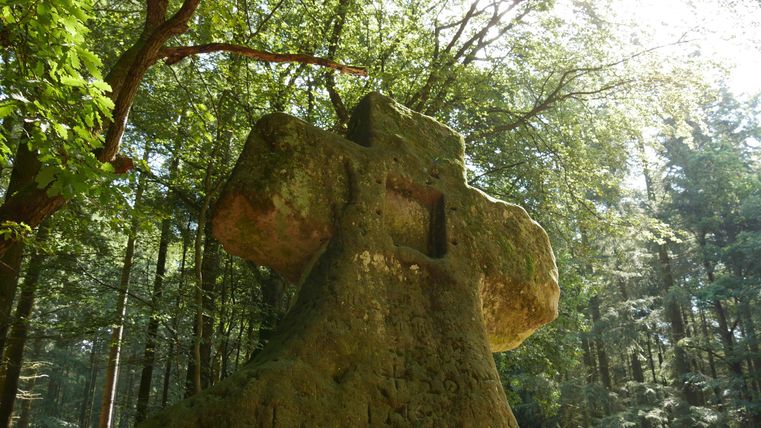 Ein großer, moosbedeckter Stein in Form eines Kreuzes steht im Wald. Sonnenlicht strahlt durch die Bäume und schafft eine friedliche Atmosphäre.