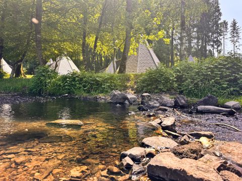 Une rivière calme avec de l'eau claire et des pierres au premier plan. À l'arrière-plan, on peut voir des tentes et des arbres luxuriants.