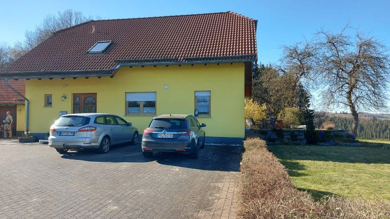 A yellow house with a red roof stands in a sunny garden. Two cars are parked in front of the house, surrounded by green lawn.
