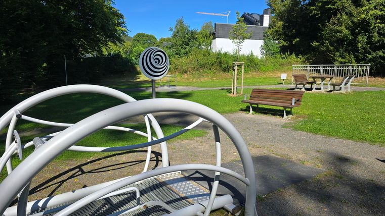 Een speeltuin met moderne apparaten en een grasveld op de achtergrond. De omgeving is groen en het is zonnig.
