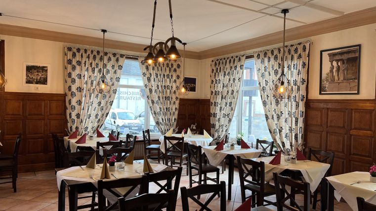 A cozy restaurant with wooden furnishings and beautifully set tables. The windows are adorned with curtains and there is warm lighting.