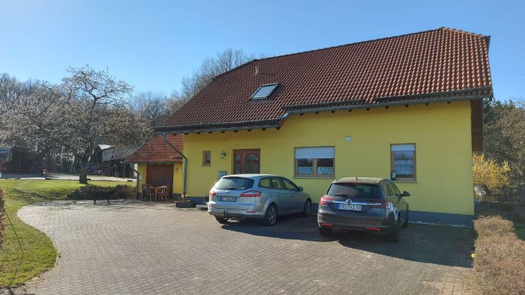 A yellow house with a red roof stands on a paved driveway. Two cars are parked in front of the building, surrounded by green countryside.