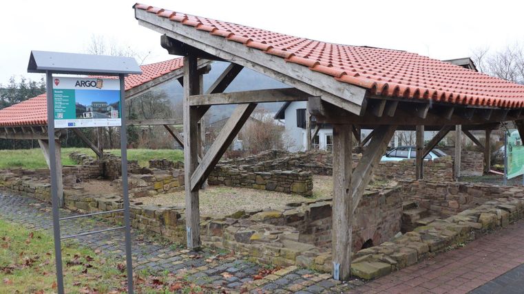 Eine historische Ruine mit einem schützenden Dach aus Holz und roten Ziegeln. Neben der Ruine steht ein Informationsschild.