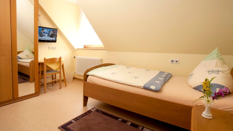 A simple room with a single bed, a television, and a mirror. The walls are painted in a light color and there is a small vase with flowers on the table.