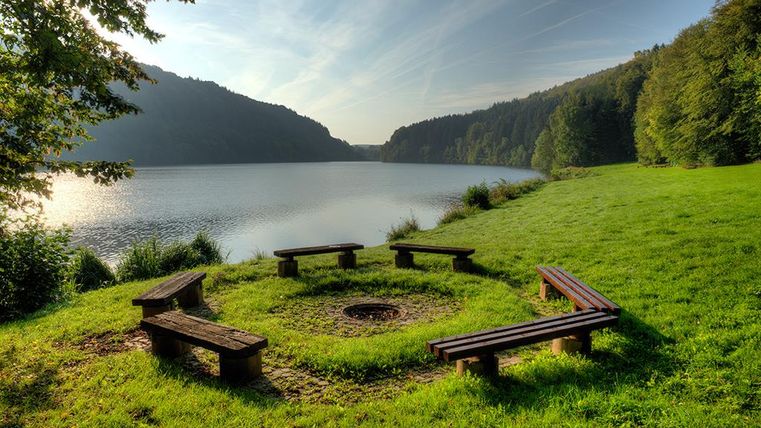 Ein ruhiger See umgeben von Wäldern. Eine Feuerstelle mit Bänken lädt zum Verweilen ein.